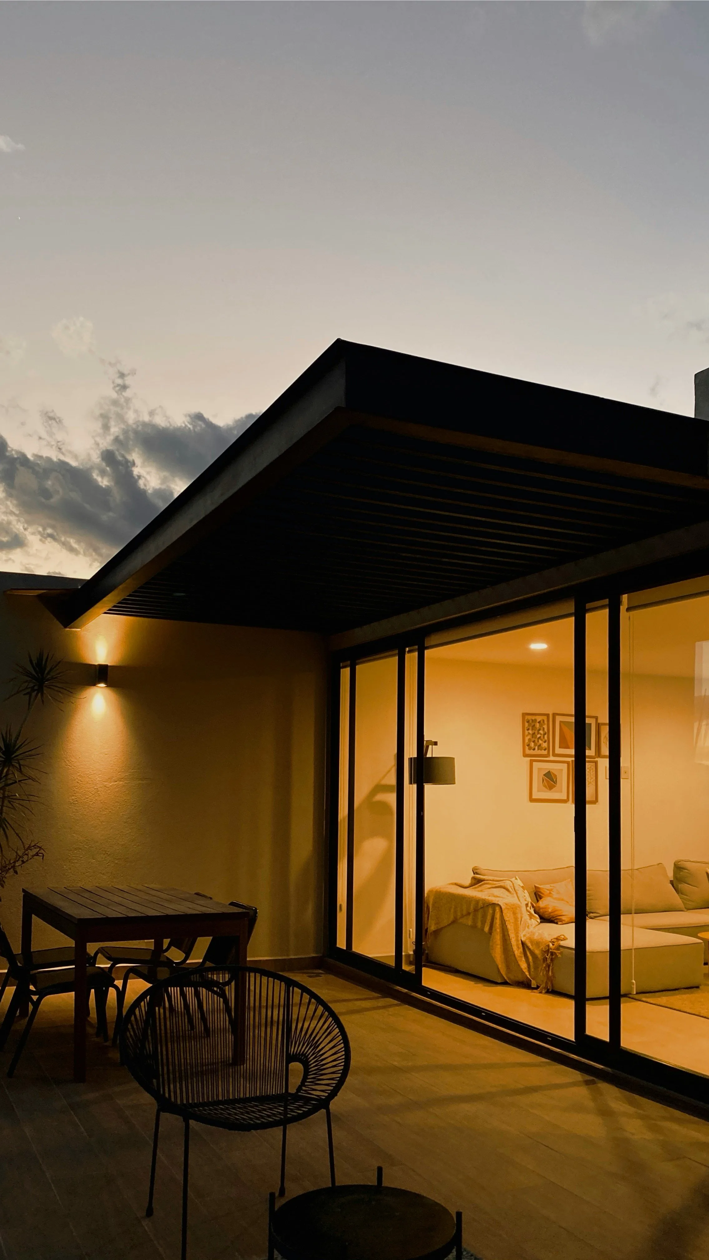 Modern single-storey home at dusk with a flat roof, full-height sliding glass doors and a deck with outdoor chairs, the interior lit warmly
