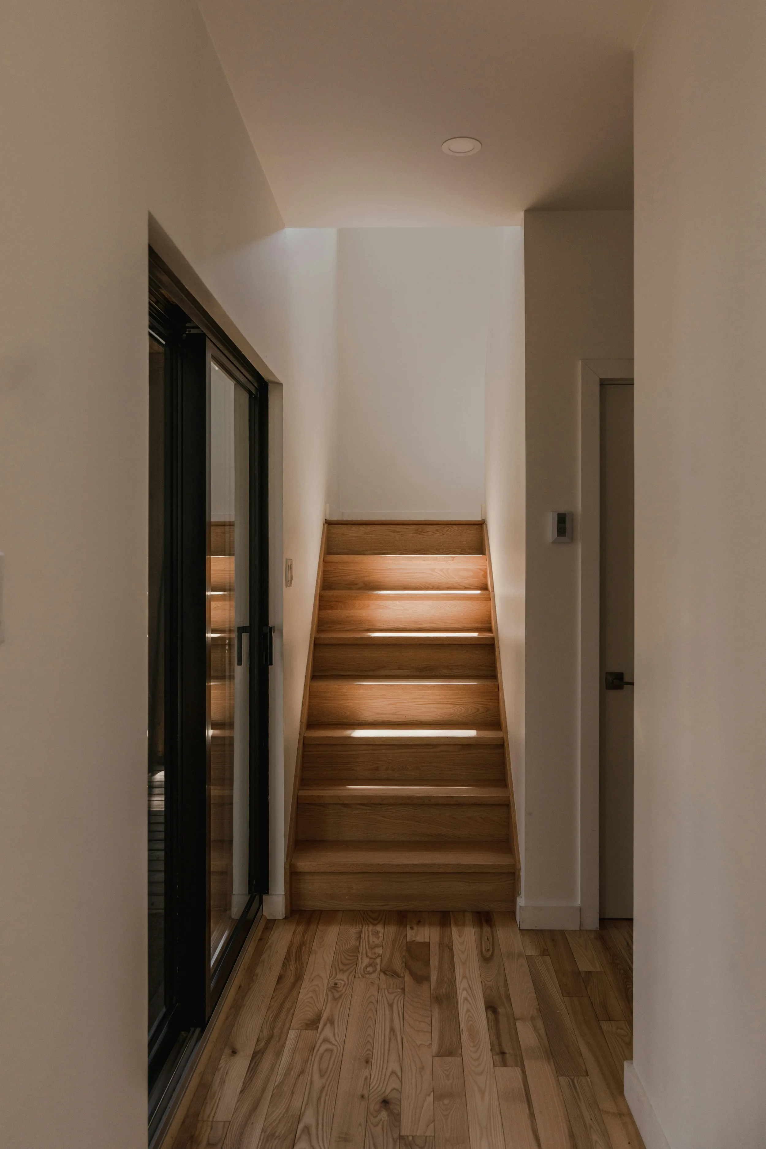 Timber staircase with lit treads rising from a white hallway with oak flooring and a glazed door to the left